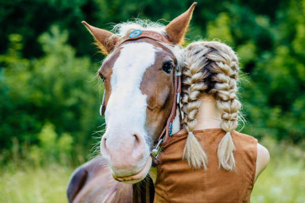 En flicka står med ryggen mot kameran och håller om sin bruna häst. Flickan har en brun väst och blonda inbakade flätor. I bakgrunden ser man lövverk och gräs.