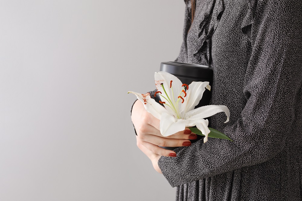 En kvinna i svart och vitprickig blus, kramar en svart urna ned askan efter sitt husdjur. I handen håller hon en vit lilja.